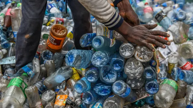 Farklı renklerdeki plastik şişeler ve bunları toplayan bir geri dönüşüm işçisinin eli. 
