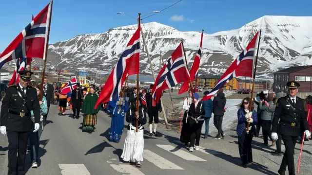 La gente en una carretera como parte de una procesión, portando banderas noruegas. Al fondo se ven algunas montañas nevadas.