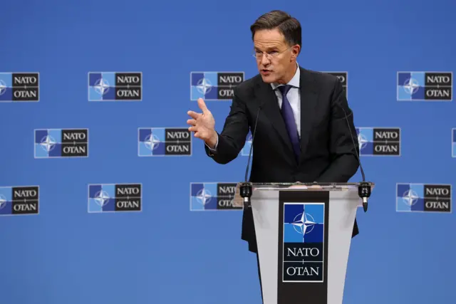 Рютте стоїть за трибуною з логотипом НАТО (синій фон із білою емблемою компаса), його права рука піднята в жесті, що виглядає як пояснення або акцент під час виступу.