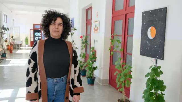 Filipa Batista, uma mulher com cabelos cacheados escuros e volumosos, vestindo jeans e camiseta preta, com um casaco com estampa de pele de vaca e mangas e gola marrons. Ela está de pé, num corredor com plantas