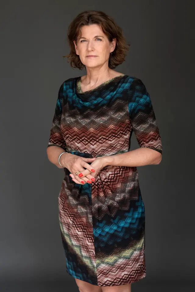 Mujer de pie frente a un fondo oscuro, vistiendo un vestido estampado de colores y con las manos entrelazadas.