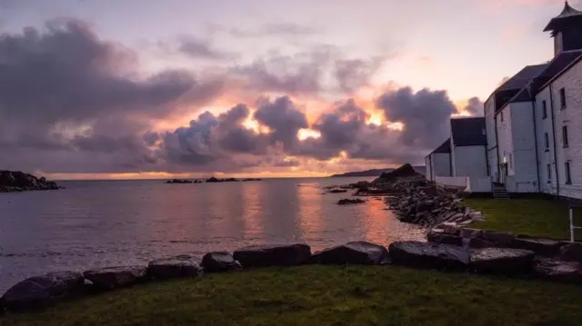 Una escena costera al atardecer con un edificio blanco a la derecha, una costa rocosa y nubes coloridas sobre aguas tranquilas.