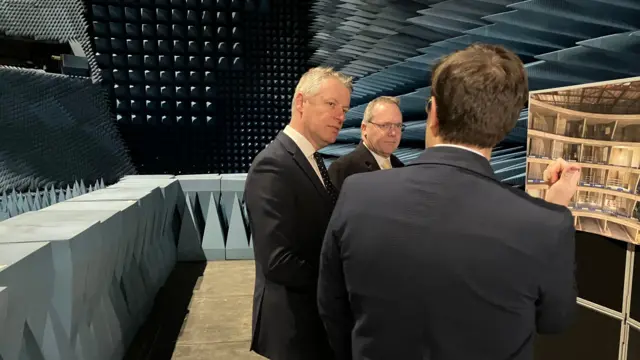 Trzech mężczyzn w garniturach rozmawia w pokoju. Ze ścian wystają duże, szare, trójkątne kolce.