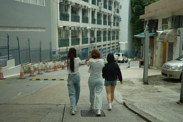 三名年輕女子從背後拍攝的畫面，她們沿著斜坡步行而下，周圍是圍欄、交通錐和建築物，呈現日常街景氛圍。