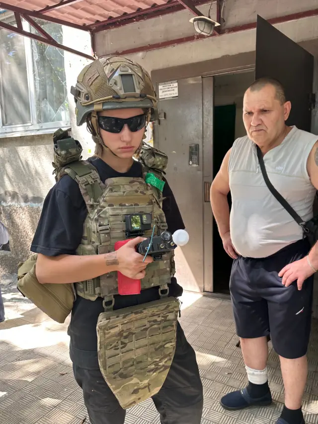 Молодая женщина Варя в камуфляжной одежде цвета хаки держит в руках детектор дронов. Рядом с ней стоит мужчина средних лет в сером жилете и синих брюках
