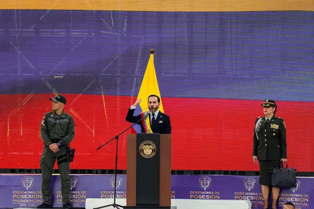 De la Espriella da su discurso de posesión sobre un escenario frente a una pantalla que muestra la bandera de Colombia y acompañado de dos representantes de la fuerza pública, un hombre y una mujer.
