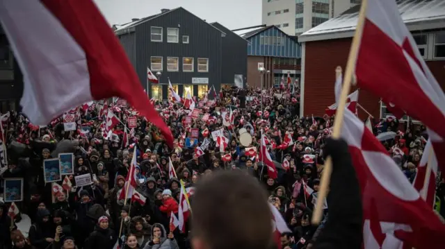 三角屋根に雪が積もった建物に囲まれた広場で、大勢が防寒具を着て、グリーンランドの旗などを持って集まっている
