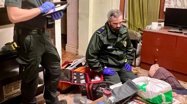 Dos hombres que trabajan en la respuesta a emergencias están de pie en una habitación, vestidos con uniforme verde de paramédico. Uno está arrodillado frente a una mujer tendida en el suelo. Su identidad está oculta, pero apenas se ven sus pies, que sobresalen de una falda a cuadros.