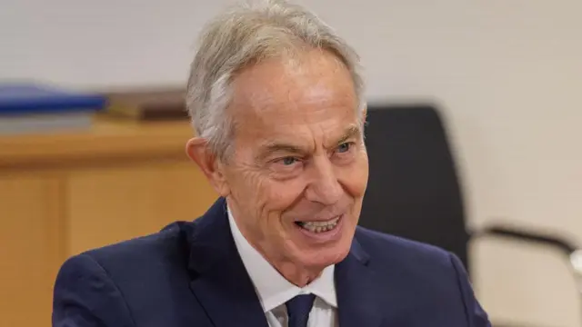 Tony Blair koyu mavi bir takım elbise giymiş. Beyaz gömlekli kravat takıyor. Kır saçlı ve arkasında tahta bir masa var.