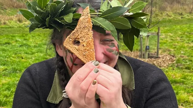 Un primer plano de Konni, sosteniendo media rebanada de pan tostado frente a un lado de su cara. Hay un pequeño agujero en la tostada y su ojo derecho mira a través de él, mientras que el izquierdo está cerrado. Sonríe con picardía. Lleva esmalte de uñas y sombra de ojos verdes, y tiene ramas pintadas en la cara.