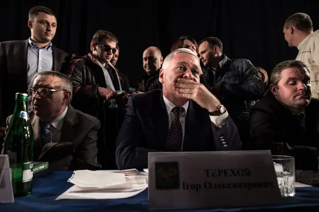 Офіційний захід у приміщенні з темним фоном. На передньому плані стіл із синьою скатертиною, на якому стоїть табличка з написом "ТЕРЕХОВ Ігор Олександрович". За столом сидить Терехов у діловому костюмі, притискаючи руку до рота. Поруч із ним і навколо кілька інших людей, які спілкуються між собою. Атмосфера виглядає напруженою.