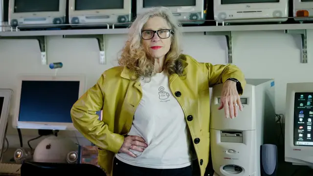 Професор Дін Грігар - жінка з довгим світлим кучерявим волоссям в окулярах та світло-зеленій шкіряній куртці