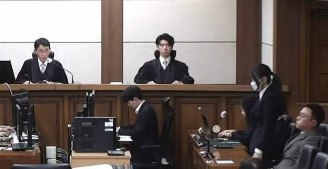 법정 내부에서 재판부와 방청석 앞에 앉은 관계자들이 보이고, 마스크를 쓴 김건희 여사가 1심 선고를 받는 모습이 보인다. 