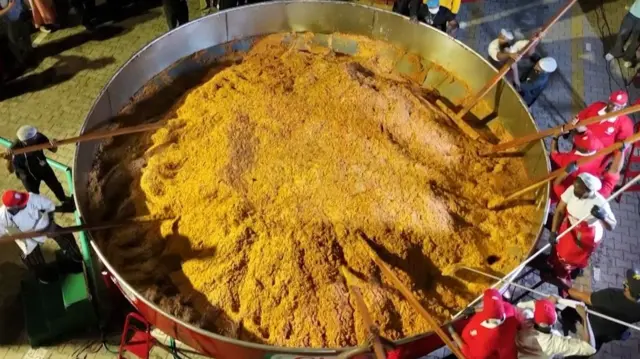 People stand around the rim of a huge pot of jollof rice and stir it using long ladles.