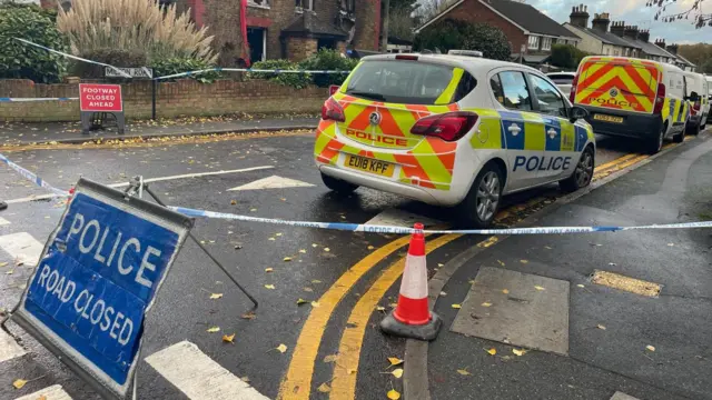 Un cartel que dice "Policía - Carretera cortada" se encuentra en el cruce frente a la casa destruida. Hay tres vehículos policiales estacionados detrás, al otro lado de la carretera.