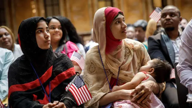 ২০১৮ সালের তেসরা জুলাই নিউইয়র্ক পাবলিক লাইব্রেরিতে আয়োজিত নাগরিকত্ব গ্রহণ অনুষ্ঠানে অপেক্ষা করছেন বাংলাদেশ থেকে আসা মোসাম্মৎ রাশেদা আক্তার (ডানে)। তিনি তার সাত মাস বয়সী কন্যা ফাহমিদাকে কোলে নিয়ে বসে আছেন। 