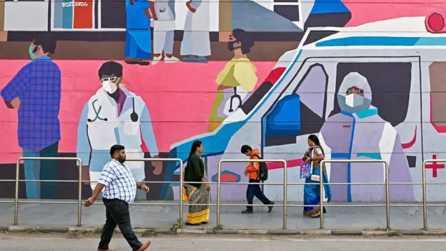 Aworan awọn eeyan to n kọja lọ lẹgbẹẹ ipolongo to n kede pe ki wọn maa lo ibomu ni India. Ọjọ Aje, ọjọ kẹfa, oṣu Kin-in-ni ọdun 2025 ni eyi. 