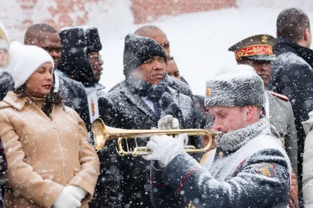 Президент Мадагаскара уже в шапке, на переднем плане трубач, засыпанный снегом. У трубача открытая меховая шапка и видно красные уши.