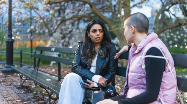 امرأتان تجلسان على مقعد بني اللون، إحداهما ذات شعر بني ترتدي جاكيت جلد أسود وقميصاً أبيض وبنطال جينز أزرق، والأخرى ذات شعر قصير جداً، ترتدي قميصاً أسود بأكمام طويلة وسترة وردية بدون أكمام.