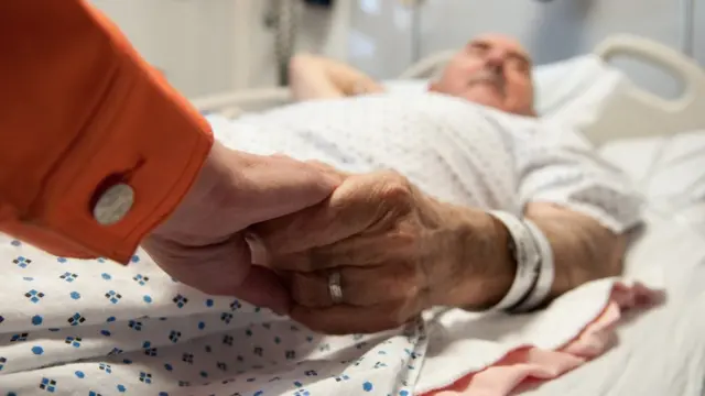 Hombre enfermo le toma la mano a su mujer.