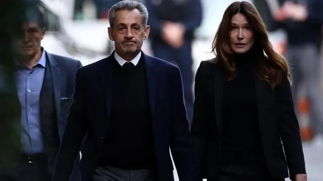 el expresidente francés Nicolas Sarkozy de la mano de su esposa, Carla Bruni-Sarkozy, salen de casa. Ambos van vestidos de color oscuro y tienen un semblante serio.