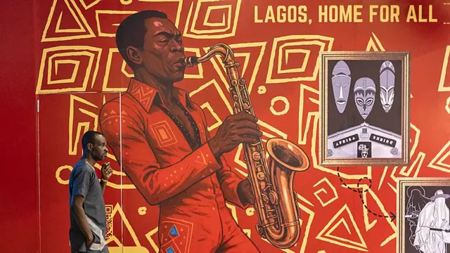 One man dey waka in front mural for Lagos, Fela Kuti wear red jumpsuit dey play saxophone, wit di words 'Lagos, Home For All'.