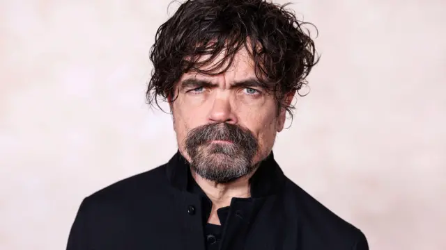 Peter Dinklage siyah bir ceket üzerindeyken kameraya bakıyor. 