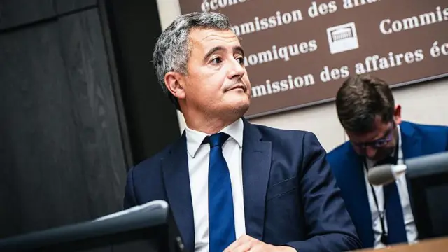 Gérald Darmanin, francuski minister sprawiedliwości, siedzi przed mikrofonem. Ma na sobie granatową marynarkę, niebieski krawat i białą koszulę.