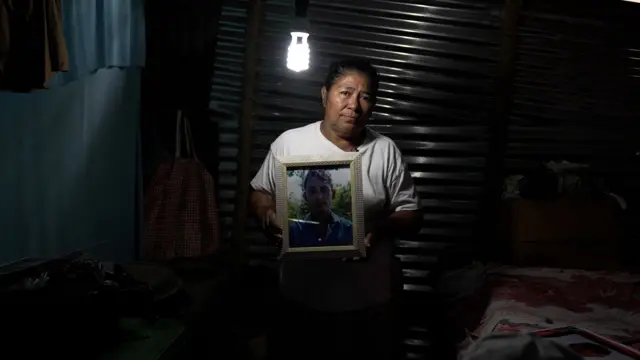 Mulher segura retrato de filho adulto; ela está num quarto de uma casa humilde, à meia luz.