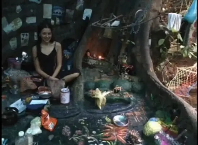 Нина Кутина в пещере рядом с корнями дерева, вокруг мелкие предметы и украшения