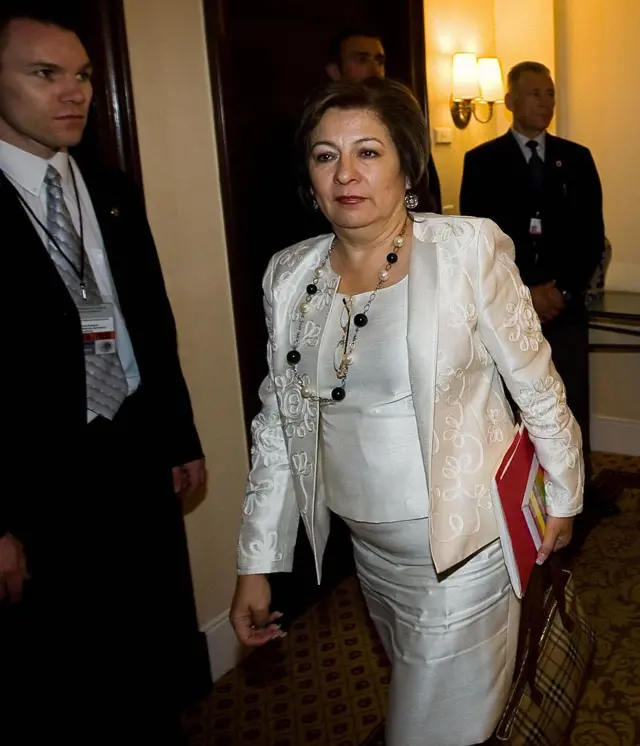 Isabel Miranda caminando en el interior de un edificio, vestida con traje de falda y chaquera, tiene un collar y lleva una carpeta bajo el brazo. Se ven dos hombres con traje y corbata en la imagen.