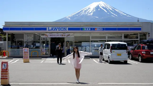 2024年5月10日，一名外国游客在日本富士山北麓的富士河口湖町，于 Lawson 河口湖站前便利店前拍照，背景可见富士山。