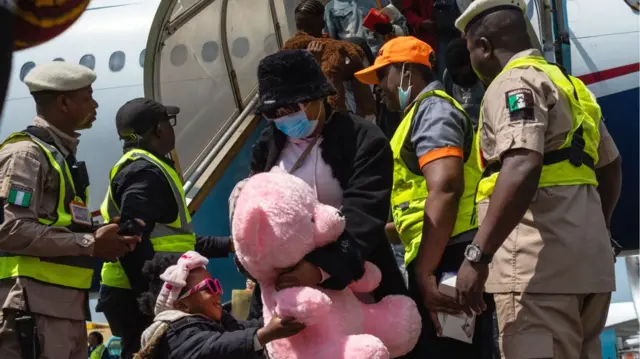 One young girl in pink sunglasses hold one giant pink teddy as she and one woman comot from inside one flight for Nigeria - security officers stand near dem