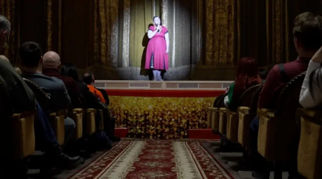 Tiyatroda bir kadın elinde mikrofonla sunum yapıyor. Fotoğraf koltuk sıralarını ayıran orta alandan ve tam karşıdan çekilmiş. Sahnedeki kadının üzerine bir ışık yansıyor ve üzerinde pembe elbise var.