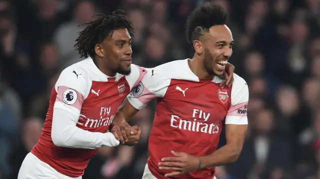 Alex Iwobi passe son bras gauche autour de l'épaule de Pierre-Emerick Aubameyang et joint ses mains à celles de son coéquipier de l'autre main, tandis que les deux joueurs célèbrent un but pour Arsenal. Ils portent tous deux le maillot domicile rouge et blanc d'Arsenal et sont filmés en plan rapproché (jusqu'à la taille), avec une foule dense floue en arrière-plan.