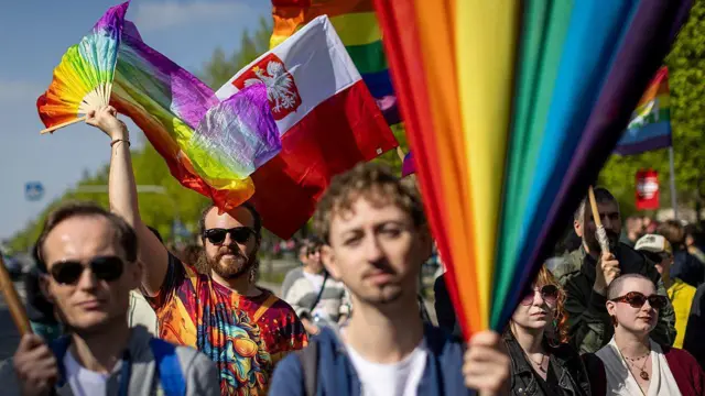 Grupa ludzi machających tęczowymi flagami LGBT i flagą polską. Mają na sobie letnie ubrania, a niektórzy noszą okulary przeciwsłoneczne. 