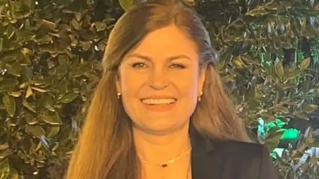 تصویری از مهرانگیز ایمن‌پور در حالی که به دوربین نگاه می‌کند و می‌خنند. پشت او برگ‌های سبز یک درخت دیده می شود