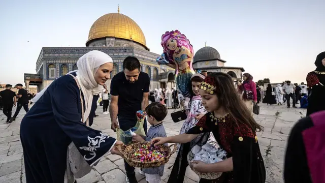 A woman dressed in black abaya wit scarf ova her head, giving sweet to young pikin while anoda man dey open nylon to give pikin sweets. Pipo dey di background