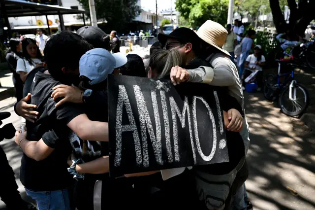 Un grupo de personas se abraza en lo que parece ser una plaza. Una de ellas tiene un cartel negro con letras en blanco que dice: ANIMO.
