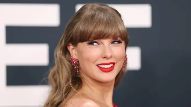 Taylor Swift, wey dey dis foto wit blonde hair, red earrings wey dey shine and red lipstick, smile ova her shoulder give di camera
