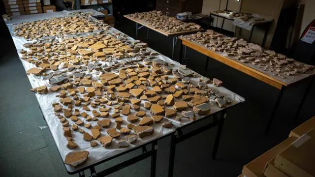 Cientos de fragmentos de yeso dispuestos sobre varias mesas. Piezas mayoritariamente amarillas que forman tres paneles cuadrados con columnas más oscuras en el medio.