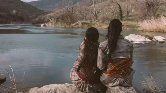 Delal (left) takes a break with a fellow fighter near a small lake close to their base. Their backs are turned to the camera and they wear traditional Kurdish dress.