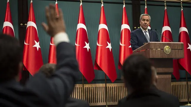 Dönemin Başbakan Yardımcısı Arınç, 29 Temmuz 2015'te bir basın toplantısında gazetecilerin sorularını yanıtlıyor. Bir gazeteci soru sormak için elini kaldırıyor. Kürsüdeki Arınç'ın arkasında birçok Türk bayrağı bulunuyor.