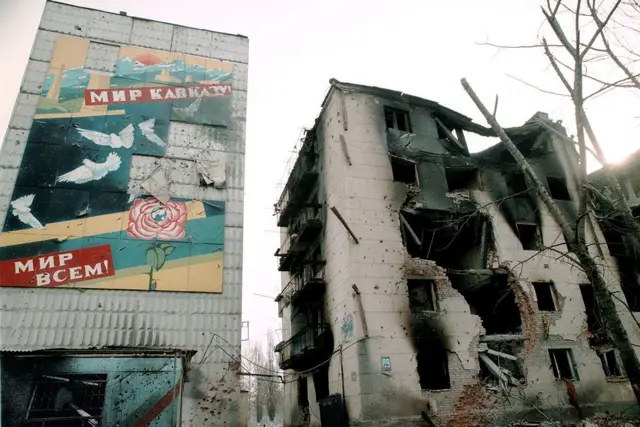 Разрушенные дома и плакат "Мир всем" в центре Грозного