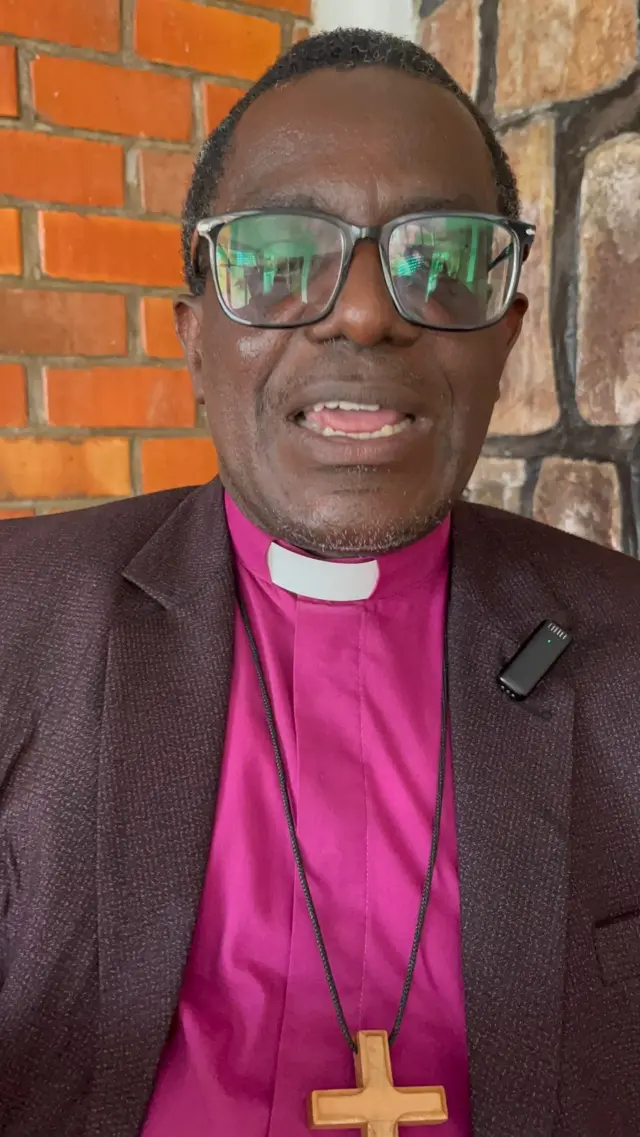 Umugabo wambaye ikositimu ijya kuba ikigina, ishati y'ibara rya roza n'umusaraba mu gatuza uri ku mugozi uri mu ijosi, arimo kuvuga