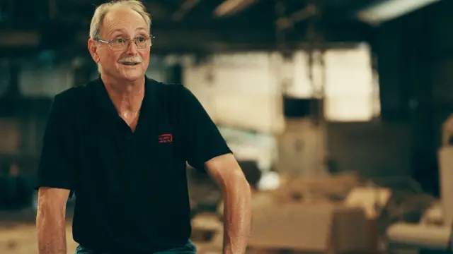 জন হার্পের চুল ছোট এবং গোঁফ ধূসর। তিনি একটি কালো ছোট হাতা শার্ট এবং সবুজ প্যান্ট পরে তার ইটের কারখানায় বসে আছেন, তার পিছনে খোলা বাক্স।