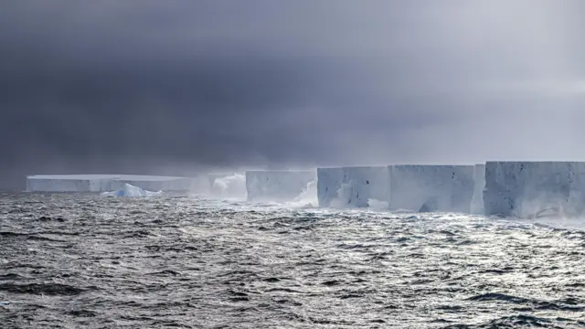 A foto mostra um iceberg enorme, com o topo plano, no mar sob um céu cinza escuro