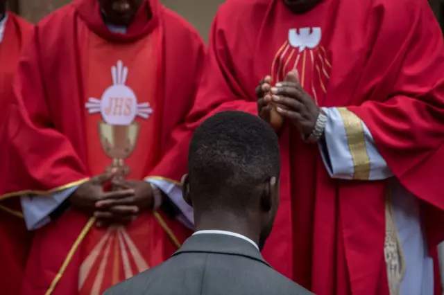Bobi Wine is seen kneeling in front of two priests.