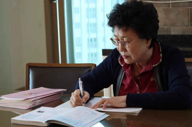 Mulher sentada lendo e escrevendo em caderno, com uma pilha de cadernos ao lado