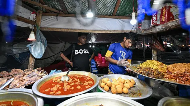 ফুটপাতের একটি দোকানে বড় গামলায় মাছ, মাংস, ডিম, ভাজাপোড়া খাবার রাখা। হাতে স্বচ্ছ পলিথিনের গ্লাভস ও নীল-হলুদ টিশার্ট পরা একজন ব্যক্তি তার পাশে কালো টিশার্ট পরা একজনকে প্যাকেটে খাবার তুলে দিচ্ছেন। 